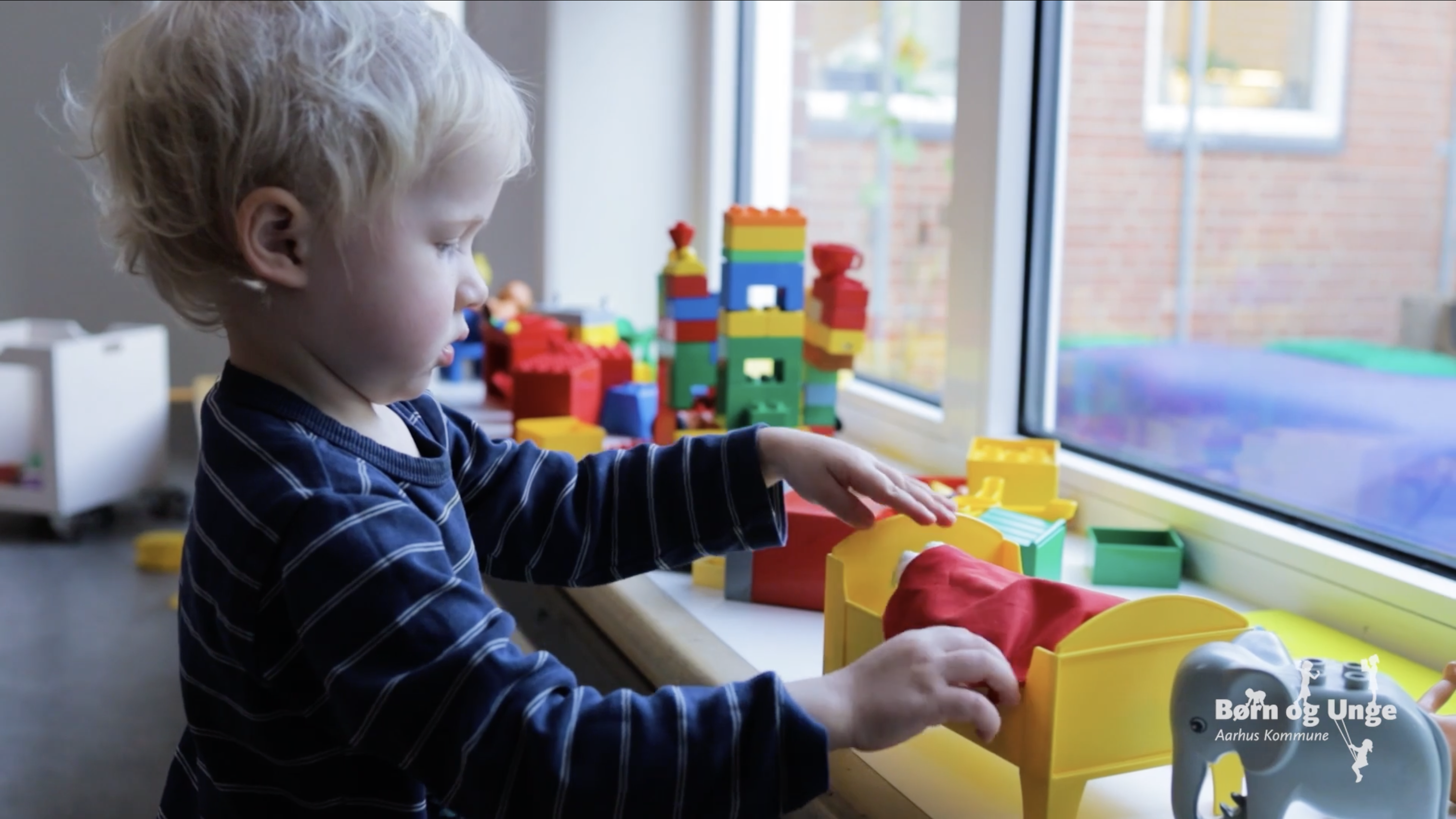 Barn leger med en dukkeseng i en vindueskarm